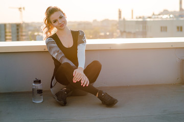 Portrat of young sporty blonde woman in stylish sportswear sitting and relaxing on the floor with plastic bottle of water after workout on the rooftop. Sunset