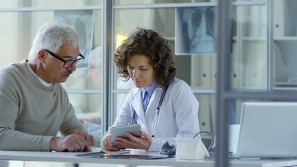 Beautiful female practitioner sitting at desk in modern doctor office with glass walls and discussing something on digital tablet with senior male patient - Powered by Adobe