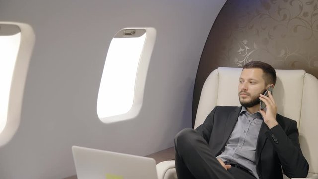 Successful person is talking on phone sitting in first class of airplane entrepreneur, bearded businessman discussing business using smartphone, looking out window in white leather chair. Concept