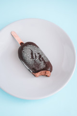 Dark chocolate bitten ice-cream, popsicle on a round plate. Pastel blue background. Minimalist food photography. Copyspace
