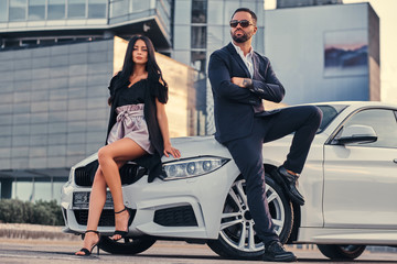 Fototapeta na wymiar Well-dressed attractive couple leaning on a luxury car outdoors against the skyscraper.