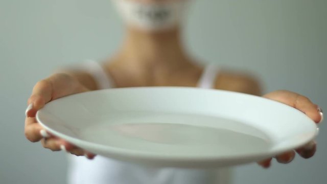 Girl with taped mouth holding empty plate, food restriction causes anorexia