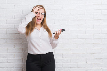 Young adult business woman over white brick wall sending message using smartphone with happy face smiling doing ok sign with hand on eye looking through fingers