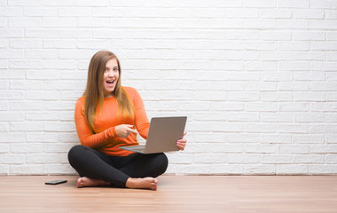 Young adult woman sitting on the floor over white brick wall using computer laptop very happy pointing with hand and finger