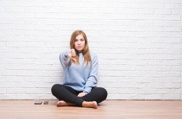 Fototapeta na wymiar Young adult woman sitting on the floor in autumn over white brick wall looking unhappy and angry showing rejection and negative with thumbs down gesture. Bad expression.