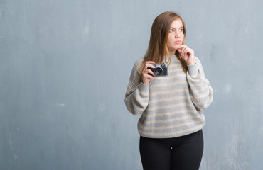 Young adult woman over grey grunge wall taking pictures using vintage camera serious face thinking...