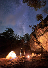 Mountain campsite at summer night amid huge rock formation. Young tourist couple, man and woman standing between small tent and burning fire under clear dark starry sky. Tourism and traveling concept.