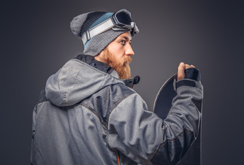 Brutal redhead snowboarder with a full beard in a winter hat and protective glasses dressed in a snowboarding coat posing with snowboard at a studio, looking away. Isolated on the gray background.