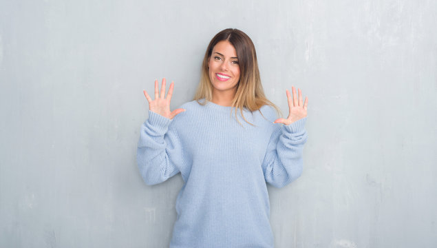 Young adult woman over grey grunge wall wearing winter outfit showing and pointing up with fingers number ten while smiling confident and happy.