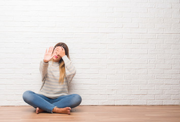 Young adult woman sitting on the floor over white brick wall at home covering eyes with hands and doing stop gesture with sad and fear expression. Embarrassed and negative concept.