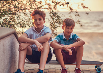Portrait of two little brothers sitting together on a skateboard against the background of the seacoast at a sunset.