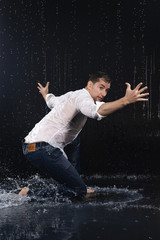 A young barefoot man wearing wet blue jeans and a white shirt expressively dances modern dances on the water under rainy waterdrops among the watersplashes