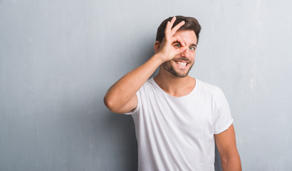 Handsome young man over grey grunge wall doing ok gesture with hand smiling, eye looking through fingers with happy face.