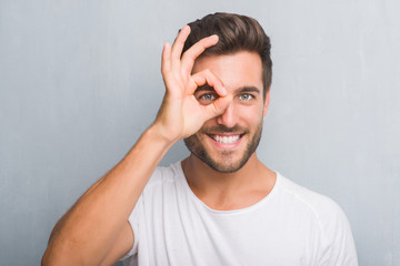 Handsome young man over grey grunge wall with happy face smiling doing ok sign with hand on eye...