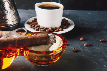 Cup of coffee, coffee beans, ashtray with cigar on dark background