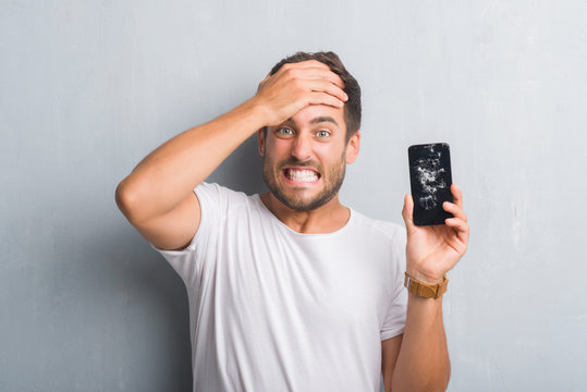 Handsome young man over grey grunge wall showing broken smartphone screen stressed with hand on head, shocked with shame and surprise face, angry and frustrated. Fear and upset for mistake.