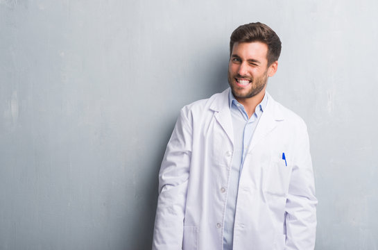 Handsome young professional man over grey grunge wall wearing white coat winking looking at the camera with sexy expression, cheerful and happy face.