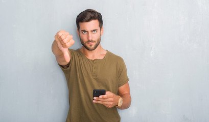Handsome young man over grey grunge wall texting a message using smartphone with angry face,...