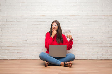 Young brunette woman sitting on the floor over white brick wall paying holding dollars serious face...