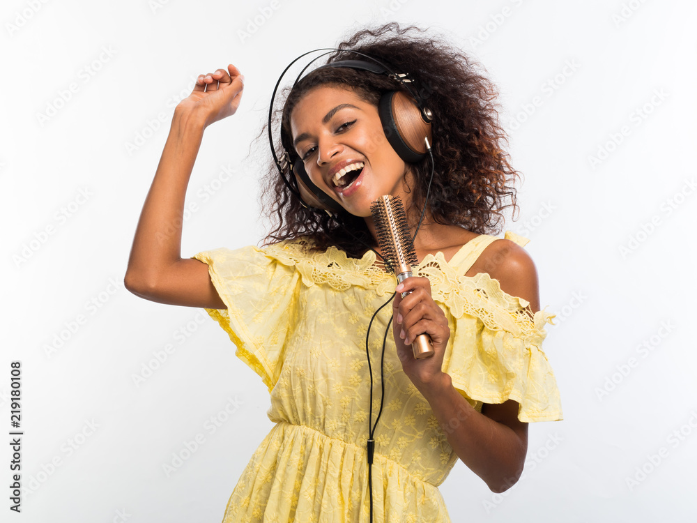 Wall mural young pretty african american woman in yellow dress singing and dancing with hair dryer instead micr