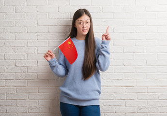 Young Chinese woman over brick wall holding flag of China surprised with an idea or question pointing finger with happy face, number one