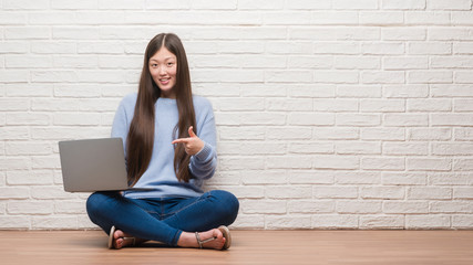 Young Chinese woman sitting on the floor over brick wall using laptop very happy pointing with hand and finger