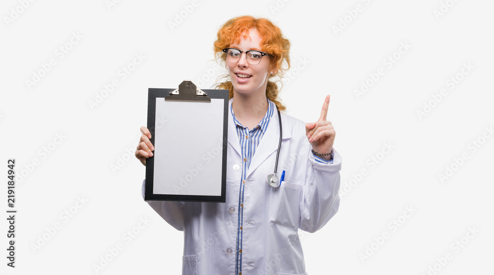 Wall mural young redhead doctor woman holding clipboard surprised with an idea or question pointing finger with