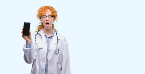 Young redhead doctor woman showing smartphone scared in shock with a surprise face, afraid and excited with fear expression