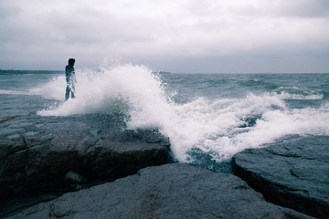 A man standing of the shore of a sea, about to be hit by a big wave.