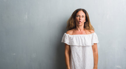 Fototapeta premium Middle age hispanic woman standing over grey grunge wall making fish face with lips, crazy and comical gesture. Funny expression.