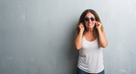 Middle age hispanic woman over grey wall wearing sunglasses covering ears with fingers with annoyed expression for the noise of loud music. Deaf concept.