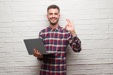 Young adult man over brick wall using laptop doing ok sign with fingers, excellent symbol