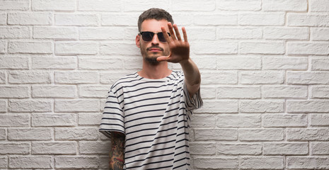 Young adult man wearing sunglasses standing over white brick wall doing stop sing with palm of the hand. Warning expression with negative and serious gesture on the face.