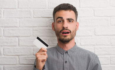 Young adult man over brick wall holding credit card scared in shock with a surprise face, afraid...