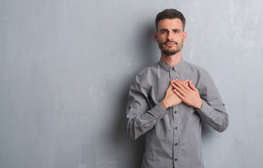 Young adult man standing over grey grunge wall smiling with hands on chest with closed eyes and...