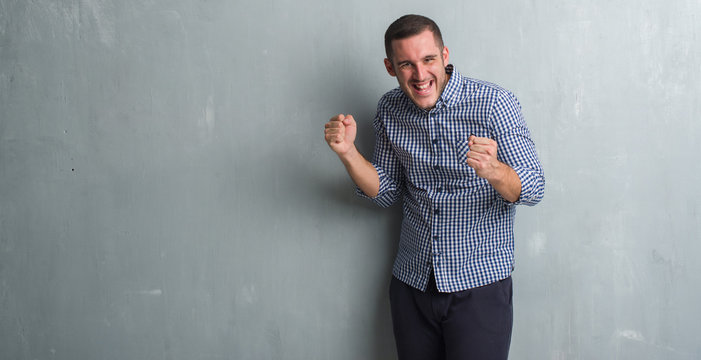 Young caucasian man over grey grunge wall very happy and excited doing winner gesture with arms raised, smiling and screaming for success. Celebration concept.