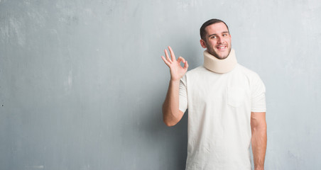 Young caucasian man injured over grey grunge wall wearing neck collar doing ok sign with fingers, excellent symbol