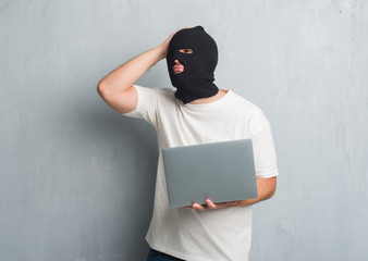 Young caucasian hacker man over grey grunge wall doing cyber attack using laptop stressed with hand on head, shocked with shame and surprise face, angry and frustrated. Fear and upset for mistake.