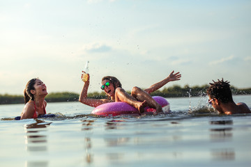 Young friends swimming and having fun in the lake.Female sitting on air mattress drinks lemonade  and having fun with friends. - obrazy, fototapety, plakaty