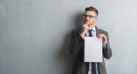 Young redhead  business man over grey grunge wall holding blank paper contract serious face...