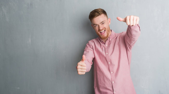 Young redhead man over grey grunge wall wearing pink shirt approving doing positive gesture with hand, thumbs up smiling and happy for success. Looking at the camera, winner gesture.