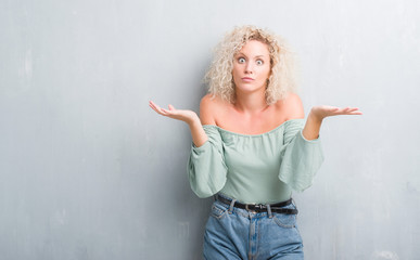 Young blonde woman with curly hair over grunge grey background clueless and confused expression...