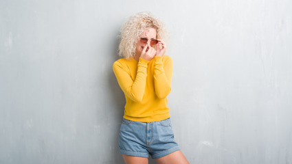 Young blonde woman with curly hair over grunge grey background smelling something stinky and disgusting, intolerable smell, holding breath with fingers on nose. Bad smells concept.