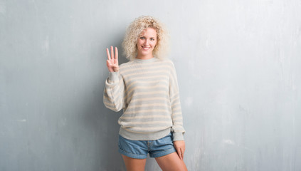 Fototapeta na wymiar Young blonde woman with curly hair over grunge grey background showing and pointing up with fingers number three while smiling confident and happy.