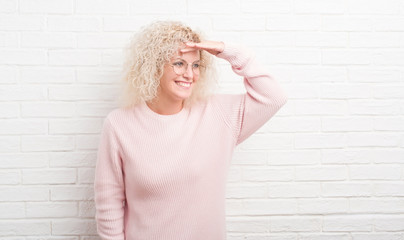 Young blonde woman with curly hair over white brick wall very happy and smiling looking far away with hand over head. Searching concept.