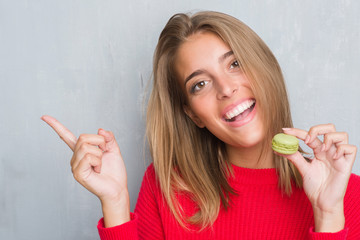 Beautiful young woman over grunge grey wall eating green macaron very happy pointing with hand and finger to the side