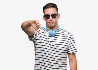 Handsome young man wearing headphones looking unhappy and angry showing rejection and negative with thumbs down gesture. Bad expression.