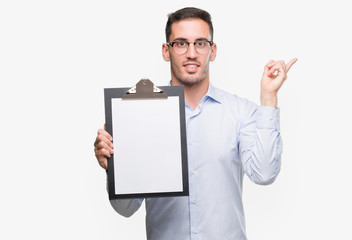 Handsome young business man showing a clipboard very happy pointing with hand and finger to the side