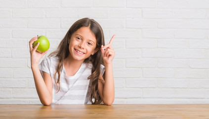Young hispanic kid sitting on the table eating fresh green apple very happy pointing with hand and finger to the side