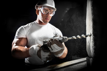 Young man using electric drill on white brick wall in a white helmet.